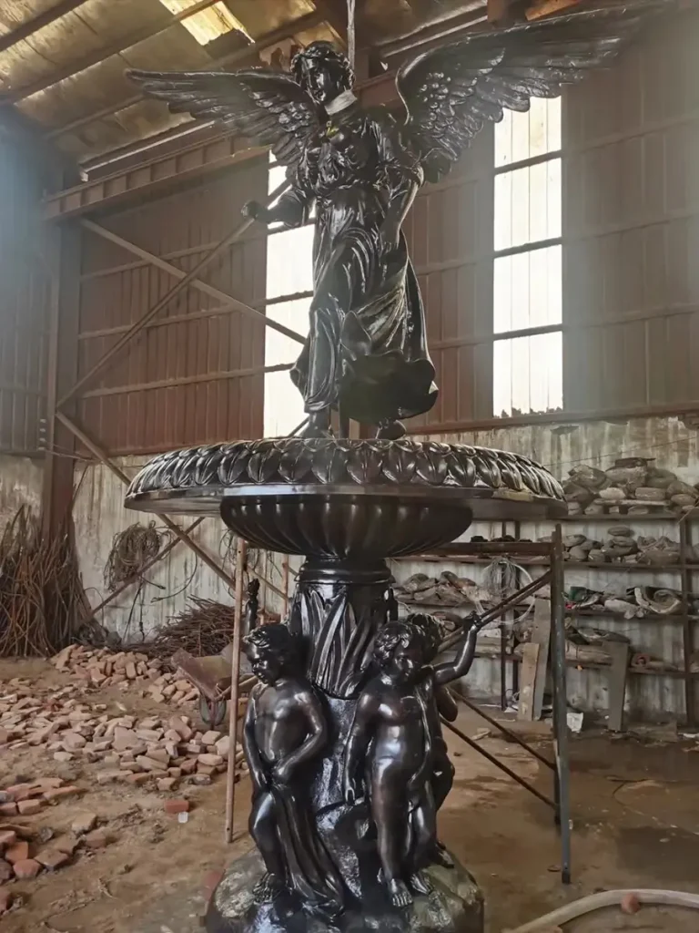 Close-up of a handcrafted bronze angel fountain statue inside the workshop, featuring a dark antique patina finish and detailed wing carving.