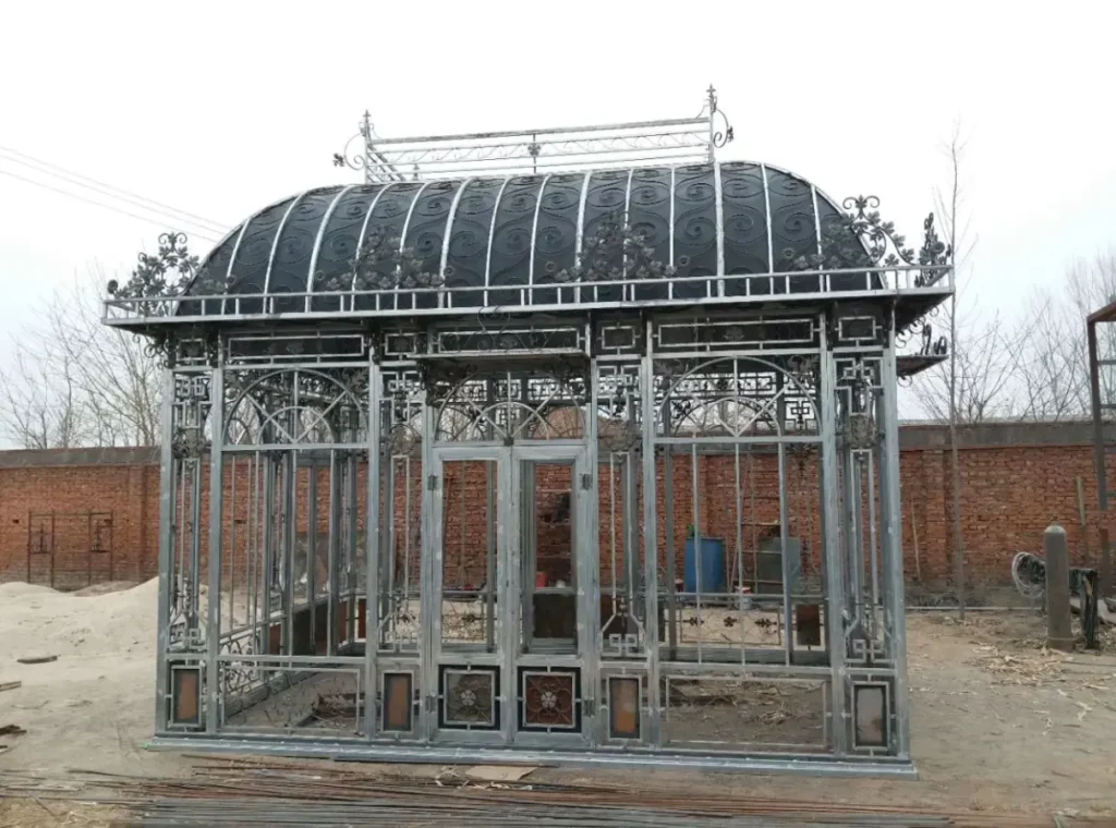 A freestanding Victorian iron orangery and glass garden house with an intricate arched roof and ornate wrought iron scrollwork.