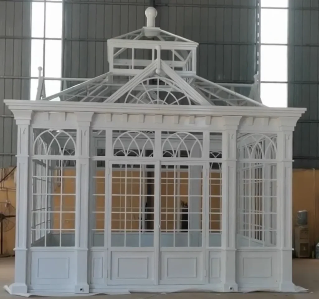 A white lightweight iron greenhouse with a fully glazed structure and tiered glass roof, designed for maximum sunlight exposure and horticulture.