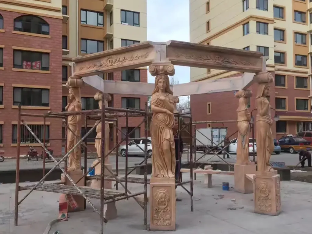 Red marble gazebo with hand-carved caryatid figure columns fully installed, showing solid stone structure before wrought iron dome placement.
