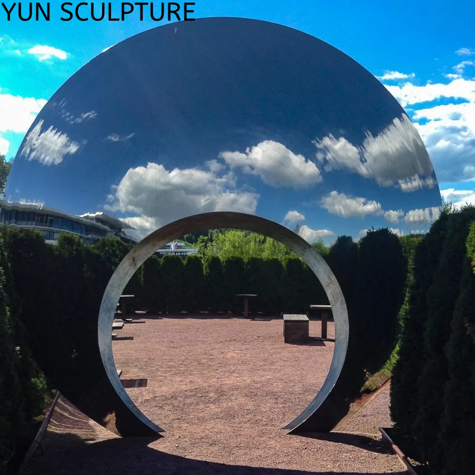 A breathtaking, monumental metal moon gate arch crafted from mirror-polished 316L stainless steel, standing across a garden pathway. Flawlessly capturing the bright blue sky and fluffy white clouds on its upper surface, this contemporary, corrosion-resistant sculpture brilliantly dissolves into the landscape, proving how the object becomes transient and almost immaterial.