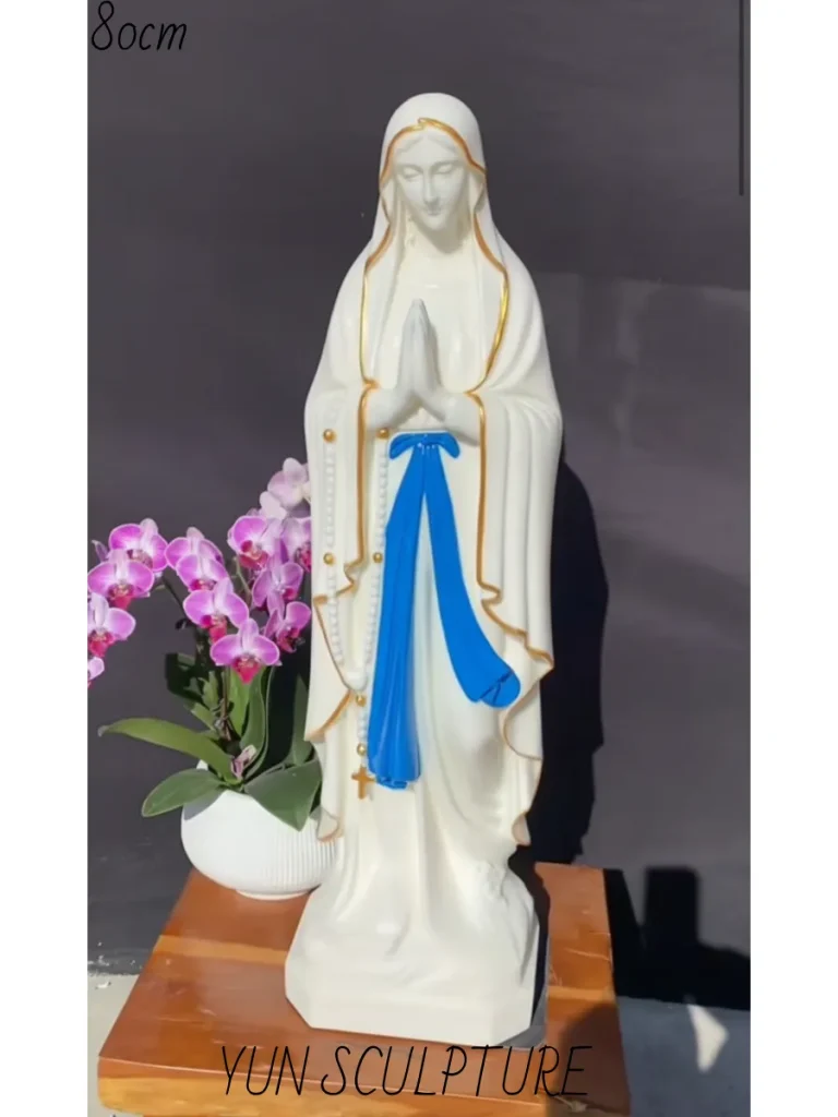 A beautifully hand-painted, 80cm tall white marble our lady of lourdes statue resting on a wooden display stand next to a potted orchid. Perfectly scaled for a small private garden or intimate courtyard, this exquisite stone figure serves as an excellent visual reference for our sizing guide, ensuring the sacred artwork feels inviting rather than oppressive when placed within personal outdoor rockwork.