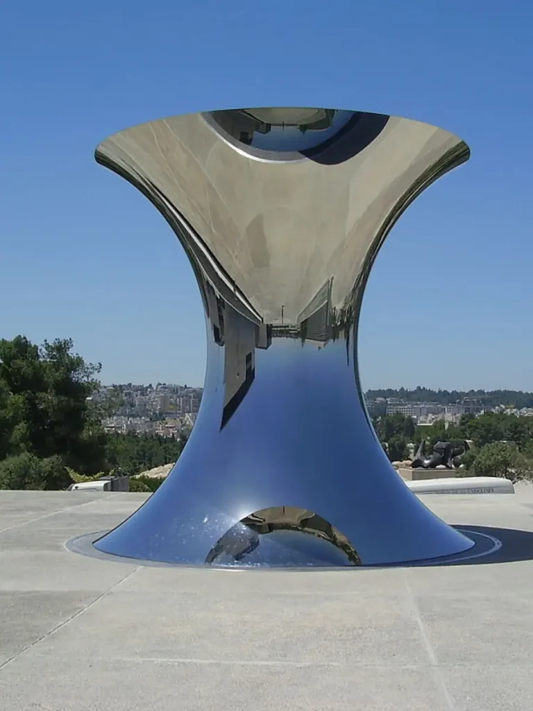 A monumental, hourglass-shaped mirror polished stainless steel sculpture standing under a clear blue sky in a public plaza. Achieving a flawless 8K mirror polish, the artwork beautifully demonstrates the "liquid silver effect" as its surface dissolves into its surroundings, capturing the sky, architecture, and ground reflections like flowing mercury, truly acting as a living mirror of the city.