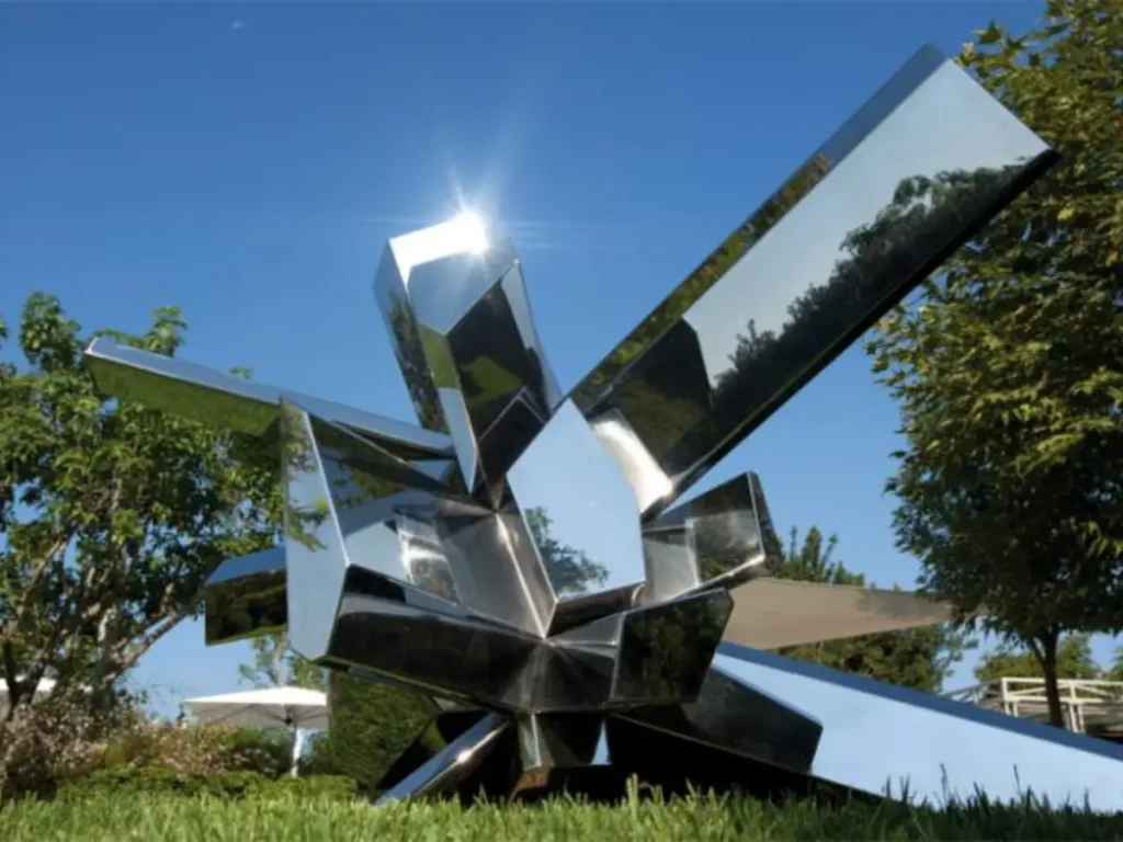 A dynamic abstract metal sculpture composed of mirror-polished intersecting planes and repeating triangular modules, resting on a green garden lawn. Capturing a brilliant sun flare on its sharp edges, this geometric artwork perfectly demonstrates how light becomes an active participant in the design, creating a powerful visual rhythm that amplifies the surrounding spatial environment.