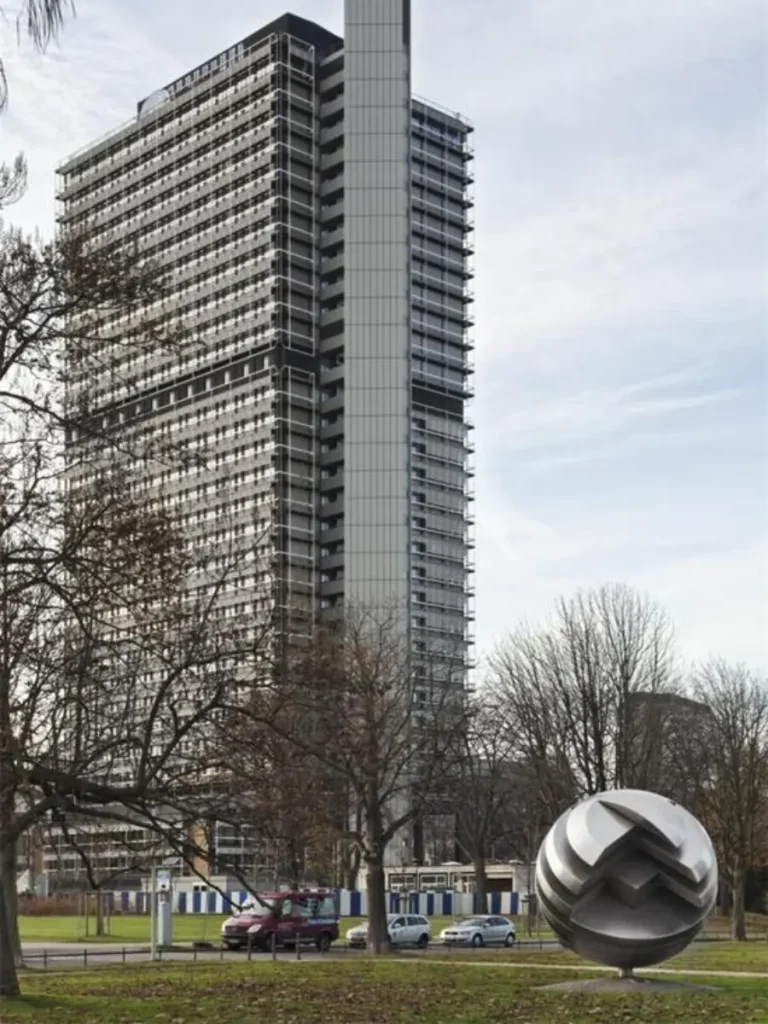 A large geometric metal sphere sculpture standing confidently on a green lawn in front of a towering contemporary skyscraper. Serving as a powerful artistic statement, this abstract metal artwork creates a memorable spatial experience that defines the corporate plaza and gives the surrounding building a distinct, unforgettable visual identity.