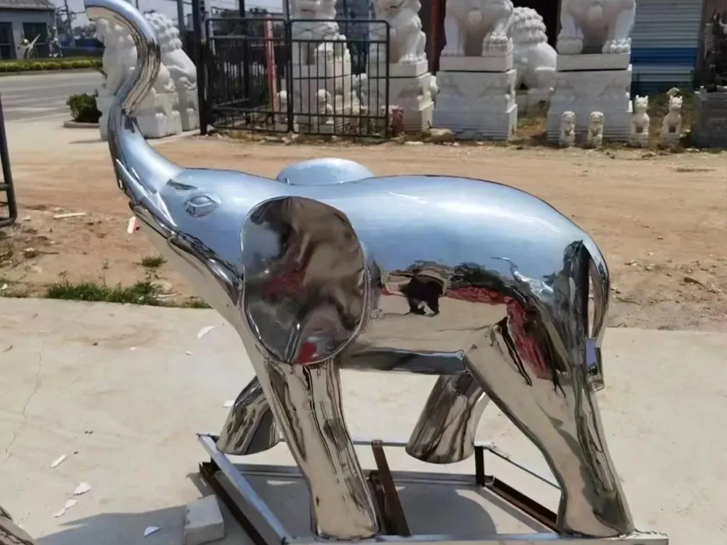 A modern abstract stainless steel elephant sculpture with its trunk raised, standing in an outdoor yard. The smooth, mirror-polished 316 stainless steel surface brilliantly reflects the surrounding sky and landscape, making it an ideal weather-resistant installation for public art spaces.