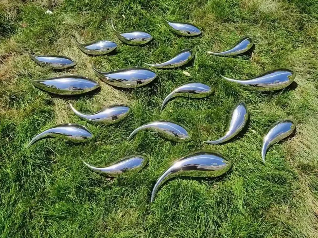 A school of abstract, teardrop-shaped mirror-polished stainless steel fish sculptures laid out on green grass. Their smooth, fluid forms catch the sunlight, ready to be installed in a garden water feature to create a mesmerizing sense of underwater movement.
