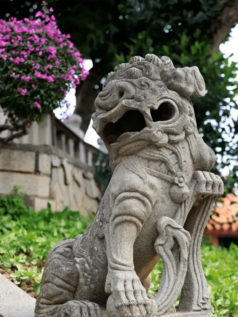 An aged, expressive stone Foo Dog in a garden setting, showcasing the vibrant coastal folk culture of Fujian. The intricately carved lion looks upwards with its mouth wide open and a flowing mane, a classic example of the agile Southern style.