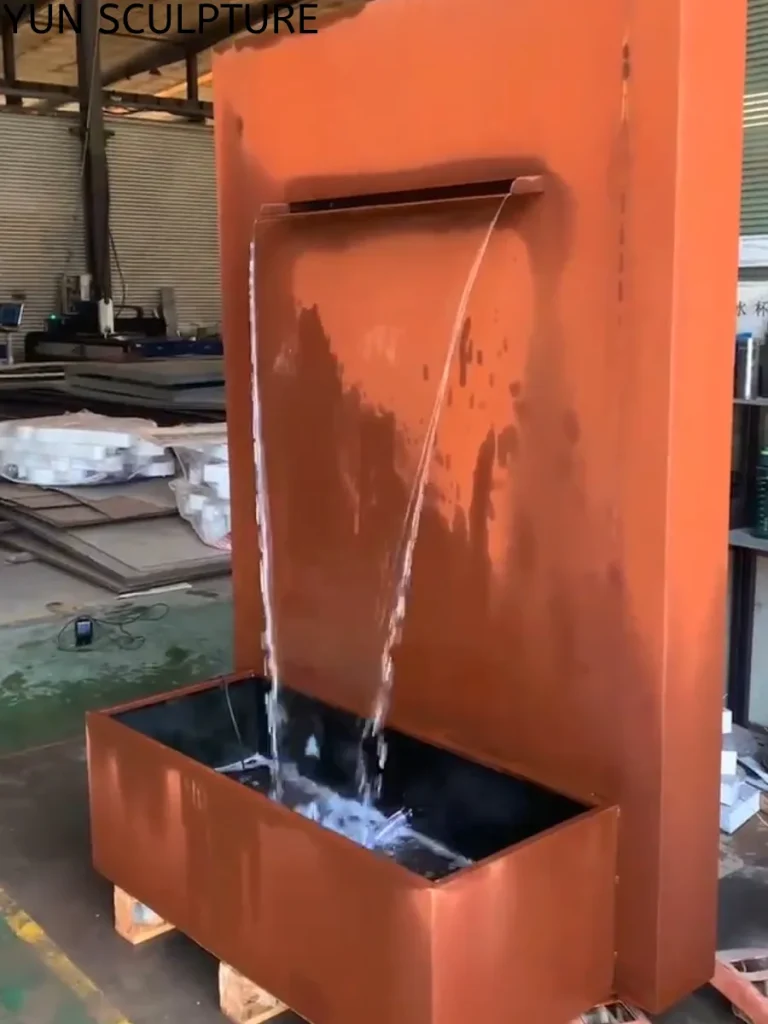 An angled perspective of a large, rusted Corten steel rain curtain water feature operating inside a workshop. By controlling the even distribution of the falling water sheet, this installation generates a consistent acoustic white noise, expertly designed to mask urban disturbance while serving as a permeable architectural boundary.