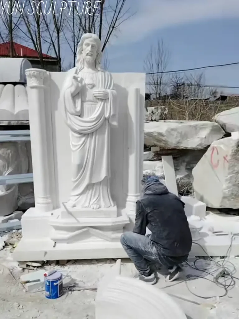A skilled artisan meticulously carving a white stone Jesus statue against a classical architectural backdrop in a workshop. This behind-the-scenes image highlights the intense precision and deep reverence required in stone chiseling to ensure every theological element—from the Crown of Thorns to the flowing robes—meets strict iconographic standards.
