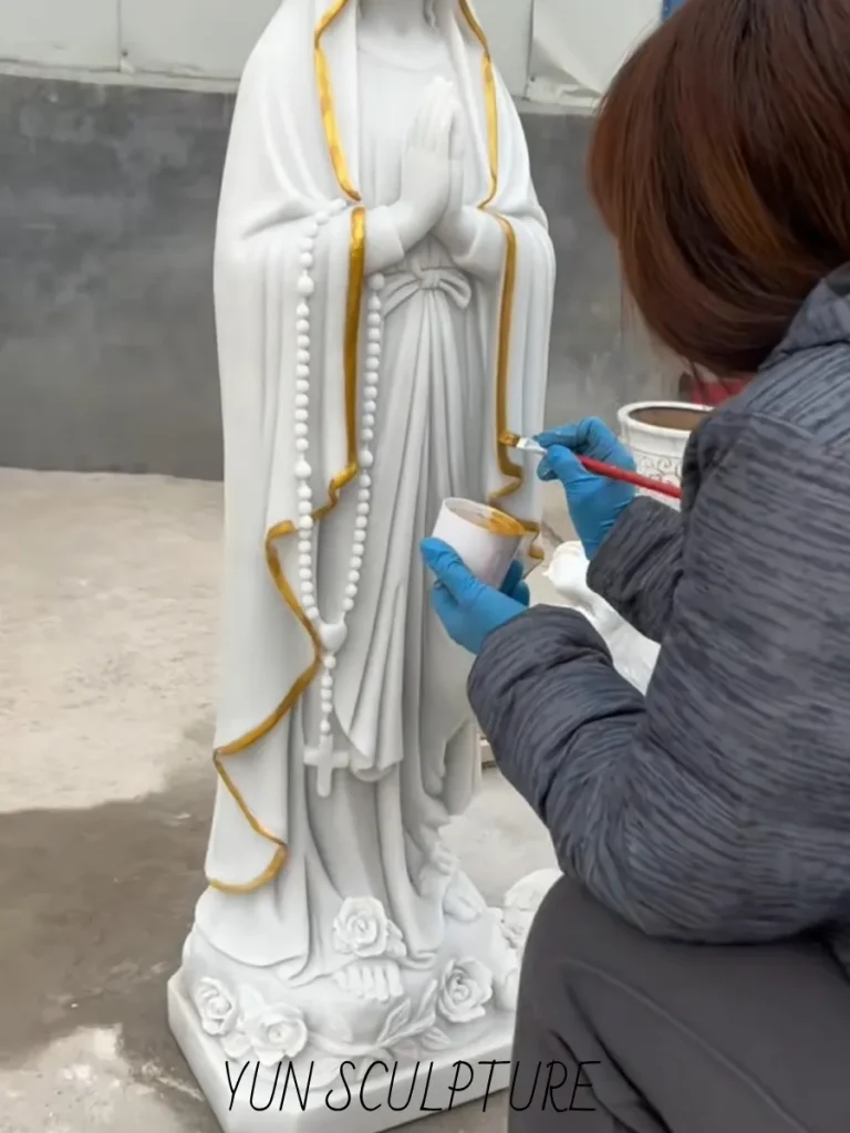 A dedicated artisan carefully applying gold detailing to the rosary and robe edges of a carved marble Our Lady of Lourdes statue. This flawless execution ensures the white beads are linked by a painted gold chain, honoring the profound theology and exact visual elements required to truly grasp the our lady of lourdes statue meaning.