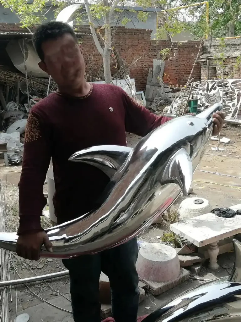 An artisan proudly holding a custom fabricated stainless steel dolphin sculpture. The flawless mirror-polished finish brilliantly reflects the worker and the factory surroundings, showcasing the premium aesthetic quality of this saltwater-safe and chlorine-resistant pool art.