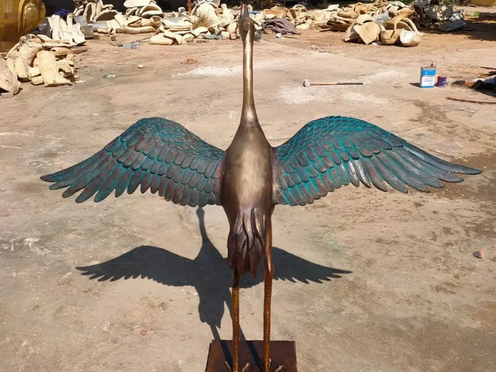 Front view of a masterful bronze crane sculpture with fully spread wings in a foundry. The artwork perfectly illustrates how bronze captures delicate feathers, enhanced by a striking blue-gray and moss green patina that adds depth and antique elegance to the heirloom piece.