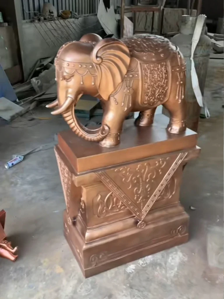 A bronze-colored elephant statue resting on an ornate pedestal with its trunk pointing down. According to cultural tradition, the trunk-down meaning reflects a quiet energy focused on accumulating resources, financial stability, and inner strength.