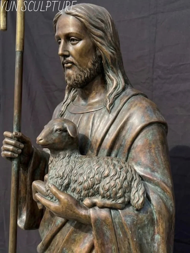 A close-up view of the serene face and upper body of a bronze Jesus statue, the Good Shepherd, holding a lamb and a staff, highlighting the detailed patina.