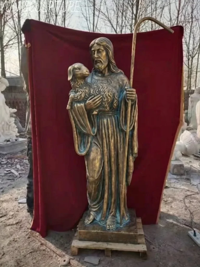 A full-body bronze statue of Jesus as the Good Shepherd, holding a staff and a lamb, standing on a transport pallet in the workshop, prepared for custom crating and international logistics.