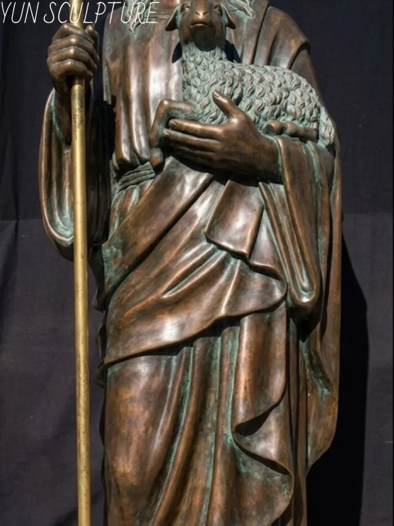 Detailed view of the torso and hands of a bronze Jesus sculpture, depicting the Good Shepherd holding a young lamb close to his chest, emphasizing the texture and patina.