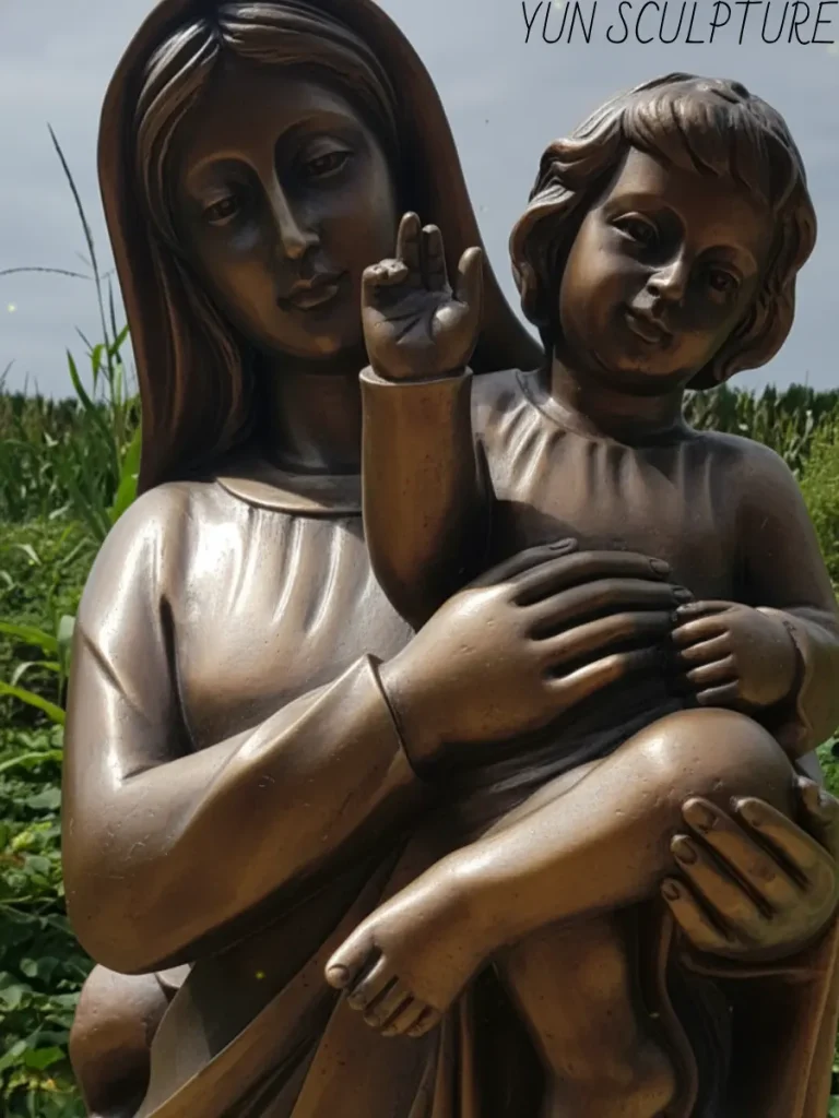 A detailed close-up of a bronze Mary holding baby Jesus statue, showing the warm statuary brown patina on the infant's skin and the refined modeling of the mother's hands.