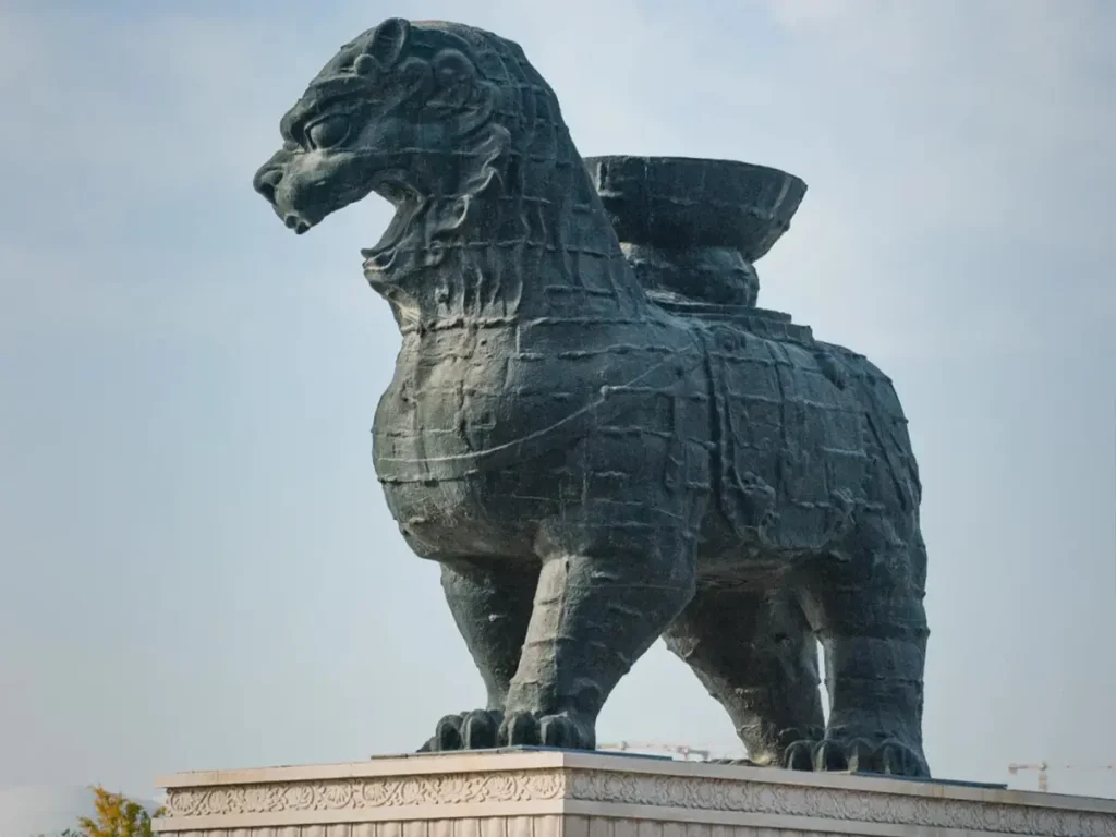 A massive cast-iron sculpture known as the Iron Lion of Cangzhou, viewed from the front-left. The dark, historic iron lion stands proudly with an open mouth on a light-colored stone pedestal, featuring a distinct lotus basin on its back against a light sky.