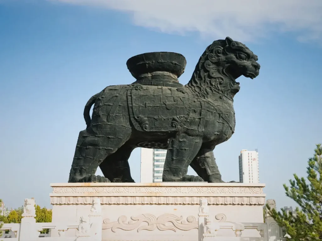 Side profile view of the historical Iron Lion of Cangzhou. The giant dark cast-iron statue stands firmly on a raised, carved stone platform with decorative railings. The image clearly displays the casting details, the large basin resting on its back, and modern city buildings in the background.