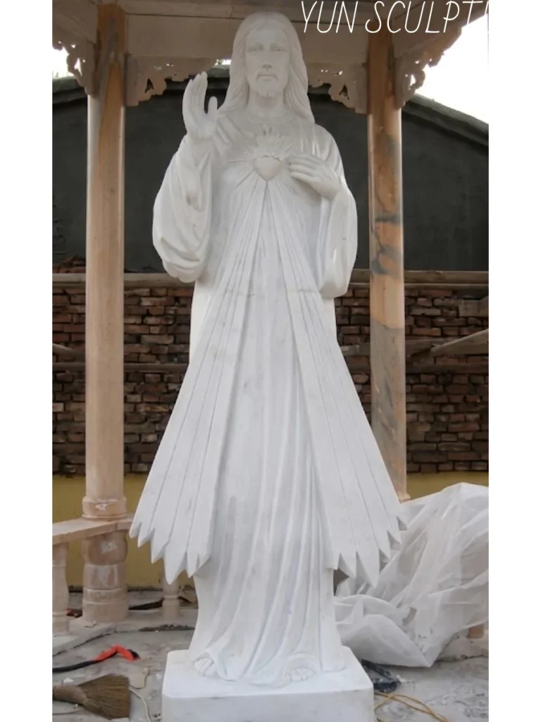 A breathtaking, finely hand-carved white marble jesus statue of the Divine Mercy image standing under a wooden pavilion in a sculptor's workshop. Perfectly demonstrating how relief and shadow replace chemical coloration in stone, the iconic rays emerging from Christ's chest are masterfully carved to capture natural translucency. Due to the delicate surface treatment, this luminous masterpiece is strictly specified for interior installation within a church or sanctuary.