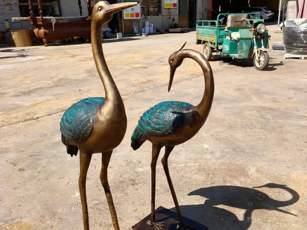 A pair of elegant bronze crane statues showcasing graceful curved necks and intricate feather details. The rich hues of the golden bronze body contrasting with the beautiful moss green patina on the wings demonstrate the evolving surface prized by art collectors.