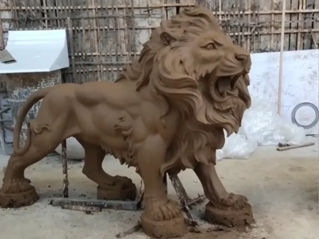 Full body view of a highly detailed clay model for a roaring bronze lion statue in a foundry. The sculptor has meticulously formed the dynamic posture and claw tension, illustrating the crucial first step of traditional casting methods for premium estate guardians.