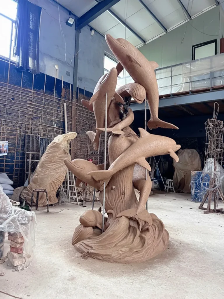 A towering clay prototype of multiple leaping dolphins resting on crashing waves inside a sculptor's workshop. This crucial design phase meticulously captures the intricate arched forms and playful rhythm, introducing pure motion into the sculpture before it is cast in bronze for homeowners searching for bespoke dolphin sculptures for sale.