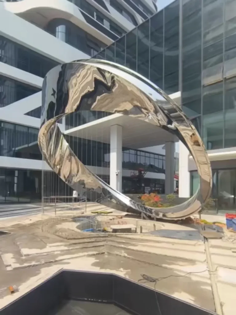 A monumental, mirror-polished stainless steel twisted ring sculpture installed in a modern corporate plaza. Thanks to the invisible engineering and internal framework, this delicate-looking geometric artwork safely transfers massive structural forces into the foundation, ensuring absolute stability against environmental elements.