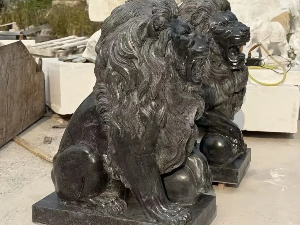 A side-angle view of a completed pair of majestic black granite sitting lion statues in a stonemason's yard, resting on pallets before shipping. These imposing sculptures feature highly detailed flowing manes and open roaring mouths, perfectly designed to establish authority and territorial control at a luxury estate entrance.