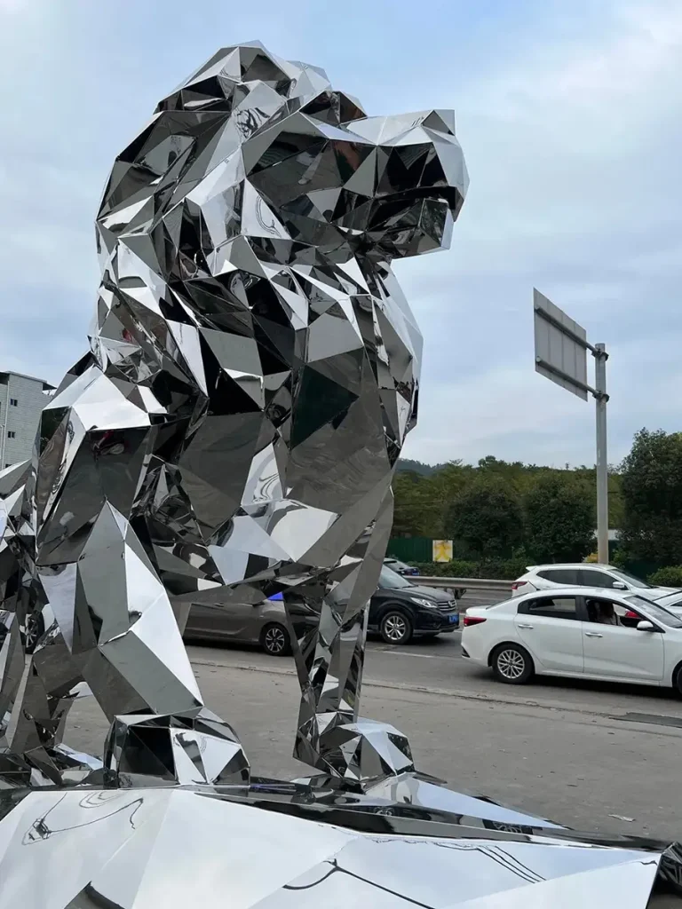 Close-up of a contemporary geometric lion sculpture featuring a brilliant mirror-polished 316 stainless steel finish. The shiny, faceted surface clearly reflects nearby cars and street signs, highlighting its striking visual impact and interaction with the surrounding environment.
