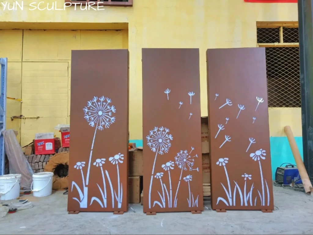 Three newly manufactured corten steel art panels featuring a dandelion design, standing in a factory ready for shipping. The varying perforation density is designed to create specific light intensity once LED systems are integrated.