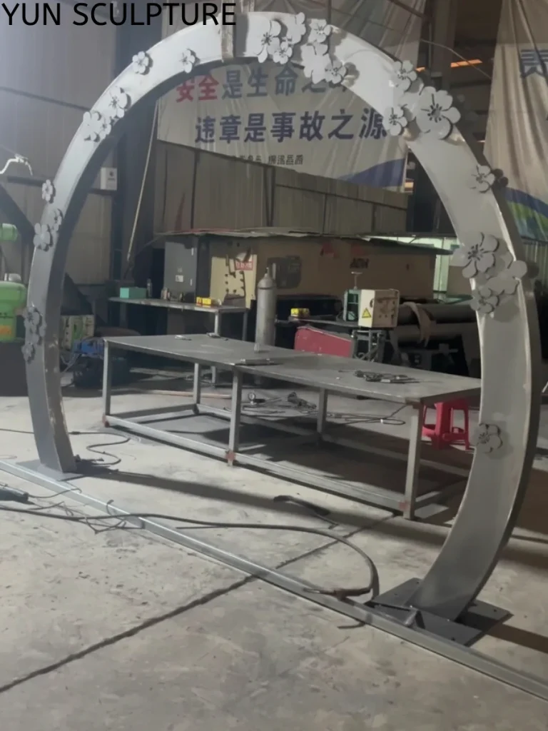 A large moon gate arch made of raw Corten steel with decorative floral details, standing in a factory workshop before the natural weathering process.