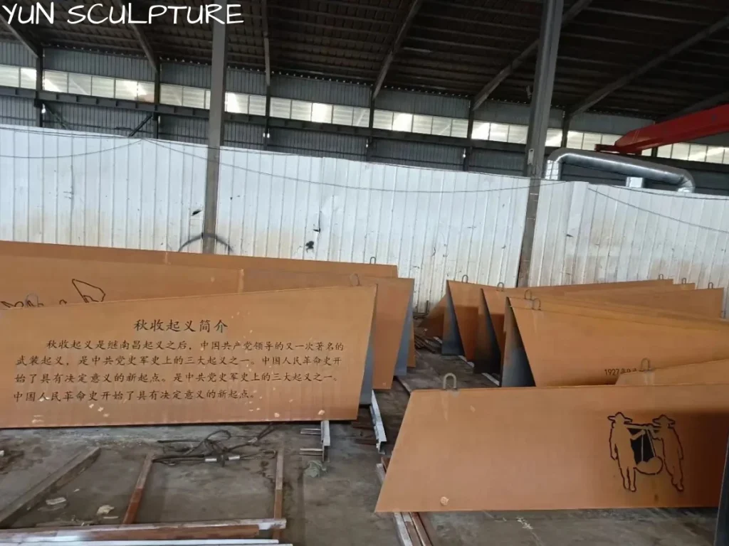 Large-format Corten steel panels with laser-cut text undergoing manual oxidation in a factory to develop a stable, protective patina for long-term outdoor durability.