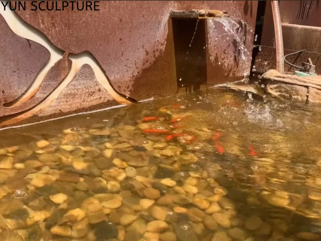 A beautiful Corten steel water feature doubling as a vibrant koi pond, with live goldfish actively swimming in crystal-clear water over smooth river rocks. This living ecosystem proves that with a fully stabilized patina or a protective Polyurea safety coating, rust runoff from weathering steel will not kill fish or disrupt delicate aquatic plants.