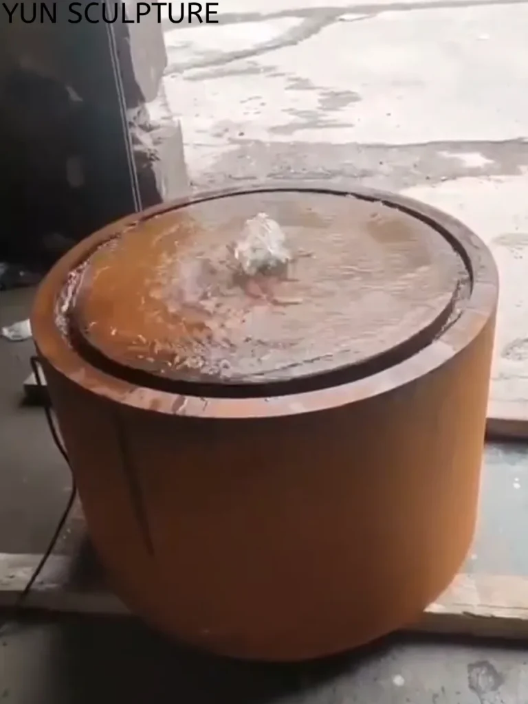 A cylindrical Corten steel water feature undergoing a fluid dynamics test in a workshop. Water bubbles from the center and forms a continuous thin film across the top plate. This expertly engineered flow pattern ensures adequate oxygen exposure, allowing the metal outdoor fountain to develop its self-protective patina without active degradation.