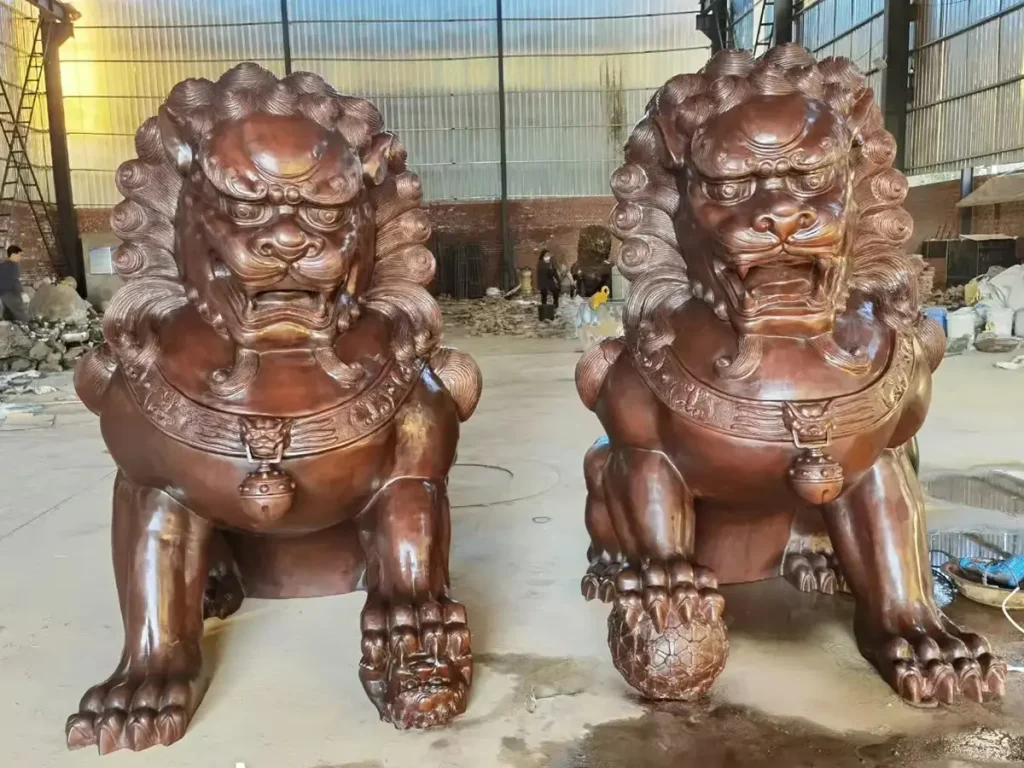 Front view of a massive pair of newly cast bronze Foo Dogs with a rich reddish-brown patina, resting on the floor of a foundry. Designed for luxury estates, these custom guardian lions feature intricate manes and chest bells, waiting to be paired with bespoke pedestals.