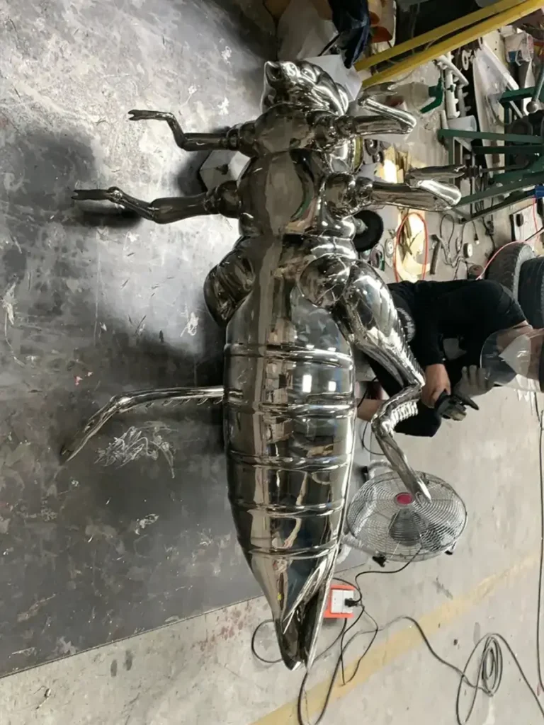 A large, mirror-polished stainless steel insect sculpture undergoing fabrication in a metal workshop. While typically considered small metal animal sculptures, dramatically scaling up these delicate creatures creates charming narrative details that invite visitors to explore.