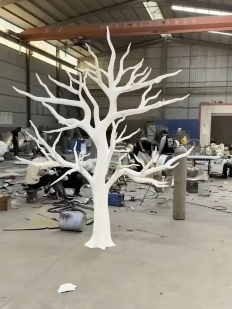 A full-scale white structural model of a tree standing in a metal workshop. This bespoke armature guides the direct metal sculpture technique, ensuring the final stainless steel tree sculpture captures a natural, non-repetitive organic form rather than relying on restrictive commercial molds.