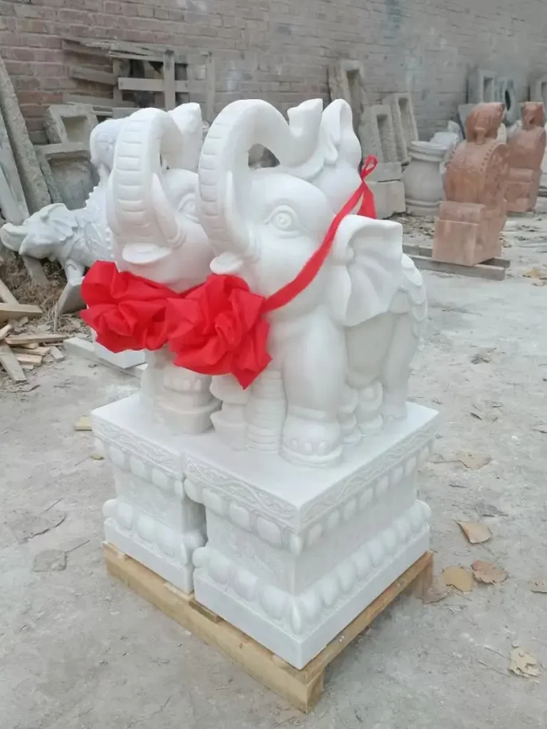 A pair of cute white marble elephant statues with their trunks raised upward, adorned with festive red ribbons in a stone workshop. In Feng Shui, this trunk-up posture symbolizes showering blessings, radiating fortune, and welcoming celebration into the home.