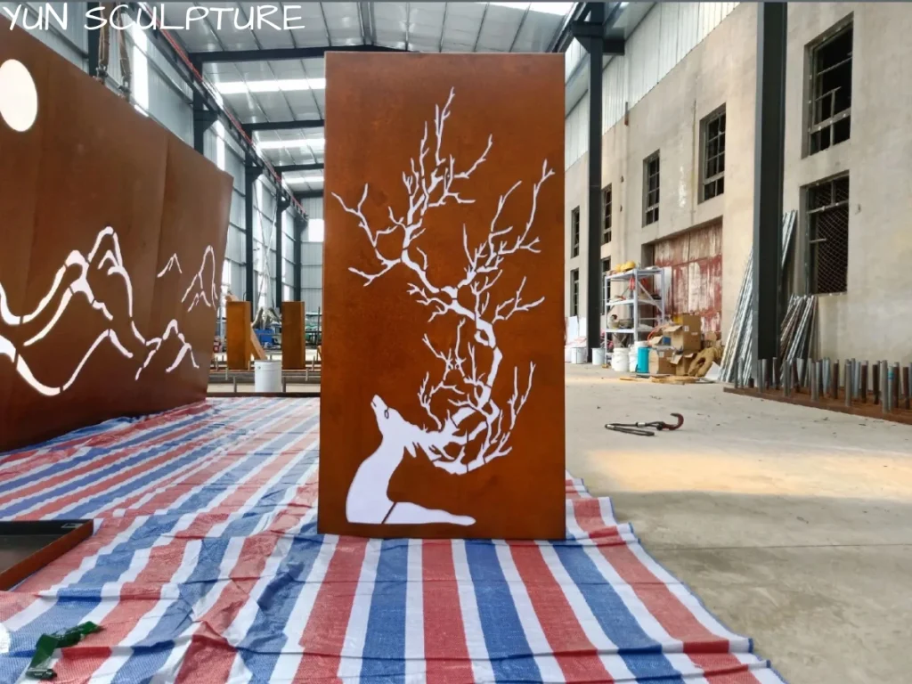 A completed laser-cut corten steel panel featuring a deer and tree motif, standing in a large factory ready for dispatch. The panel is engineered to become a luminous sculpture once LED strips are mounted within its rear cavity.