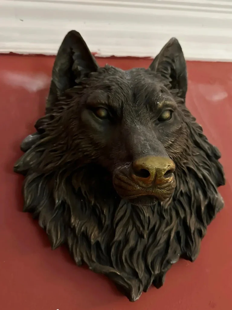 Close-up of a high-quality bronze wolf head sculpture mounted on a reddish wall. The exceptional craftsmanship is visible in the crisp, deep textures of the fur, demonstrating how true cast bronze retains its fine details instead of fading like mass-produced concrete.