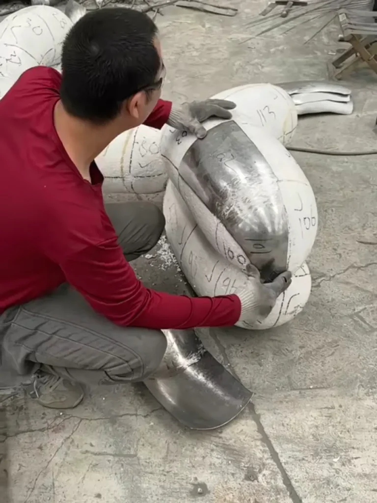 A skilled craftsman carefully fitting a hand-hammered stainless steel panel onto the foam structural mold. This demonstrates the direct forging and shaping technique, ensuring the metal perfectly matches the intended sculptural form while remaining hollow inside.

