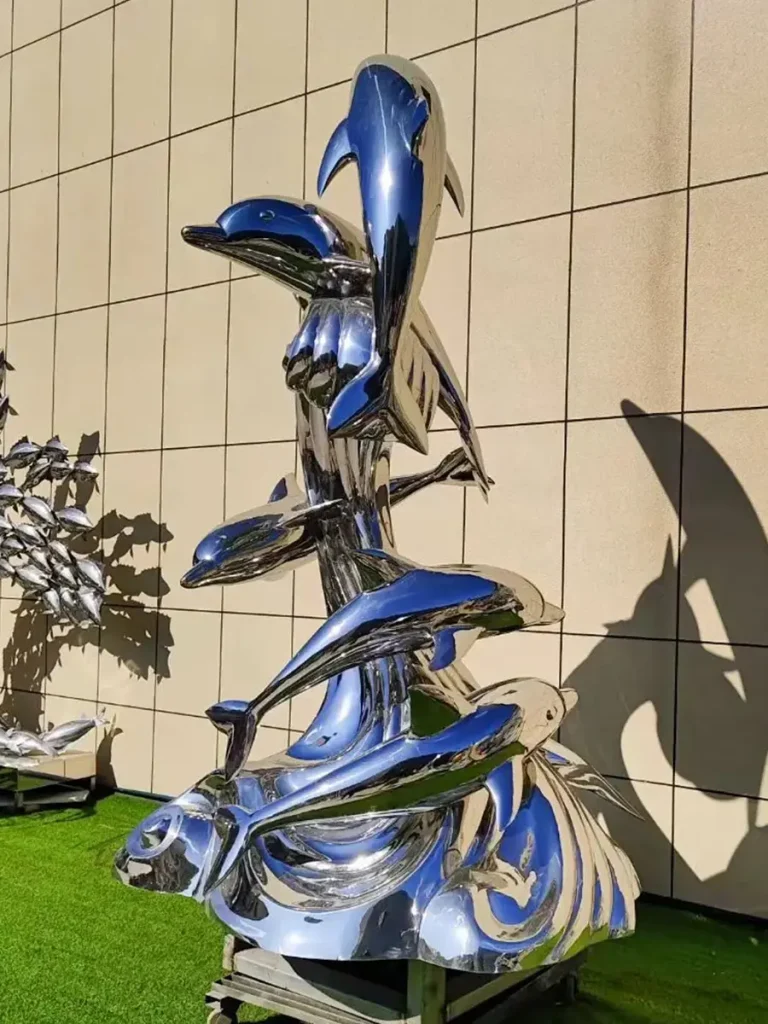 A towering, dramatic installation featuring multiple mirror-polished stainless steel dolphins leaping from a sculpted metal wave. The highly reflective surface amplifies the illusion of marine life emerging directly from the water under the sunlight.