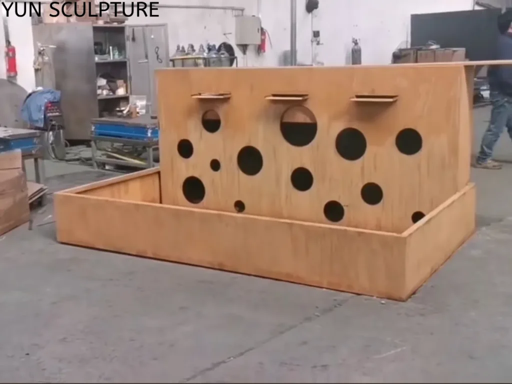 A newly fabricated Corten steel water wall and planter basin resting dry on a factory floor. Featuring precise geometric circular cutouts and three spillway spouts, this raw metal framework showcases the stark industrial character and engineered structure required to securely support living ecological systems in modern landscapes.