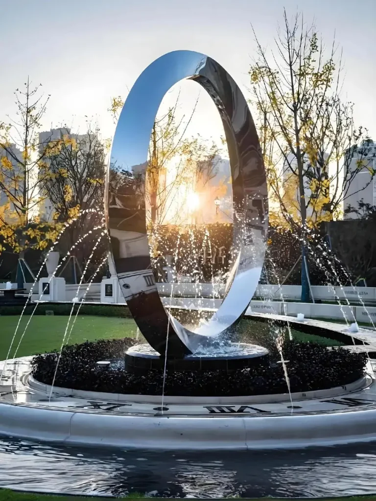 A massive mirror-polished stainless steel ring fountain standing securely in an outdoor water feature. The sunset reflects beautifully off the metal surface while water jets spray around it. This seemingly effortless display relies on hidden pre-embedded structural anchors and waterproof gaskets to safely support the multi-ton heavy metal art.