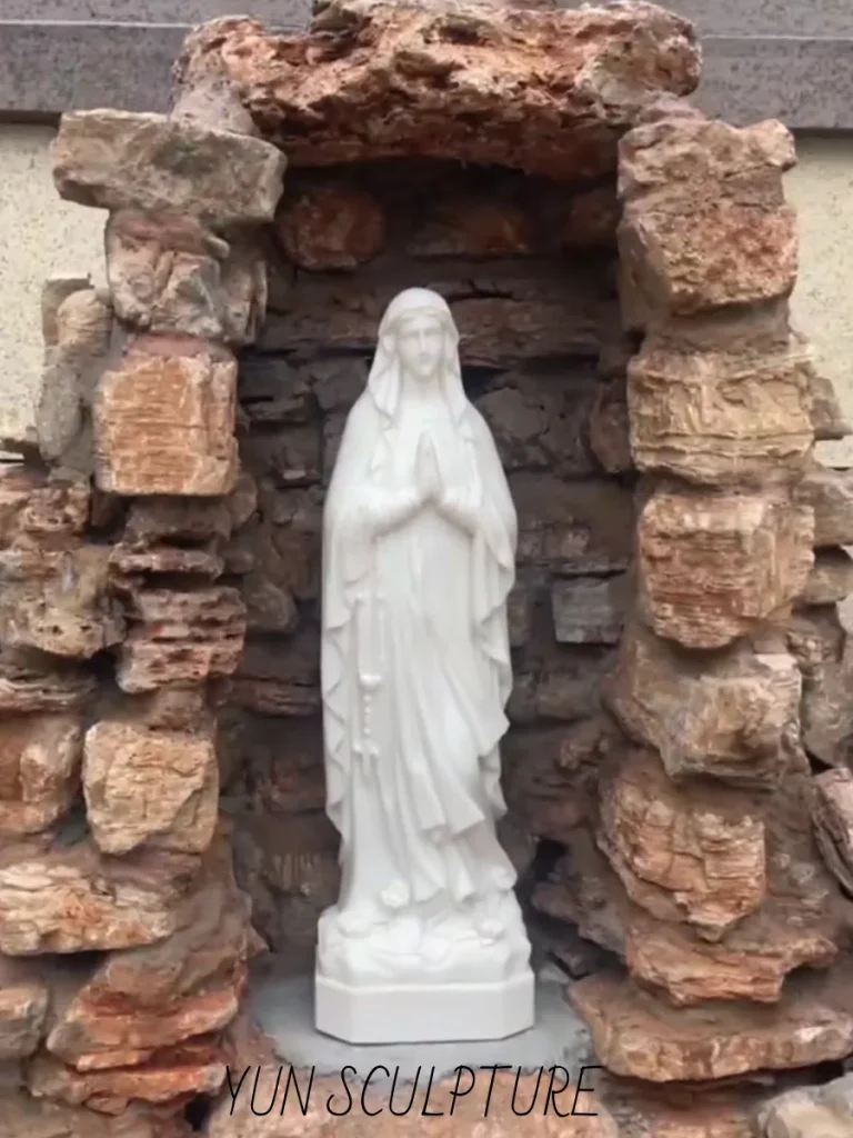 A full frontal view of a partially constructed, custom-built stone cavern framing a delicate white marble Marian statue. Showcasing the intense structural engineering behind every complete our lady of lourdes grotto garden statue landscape, this robust, arched masonry structure acts as a crypta, specifically designed to elevate the primary theological focal point and permanently shelter the sacred artwork.