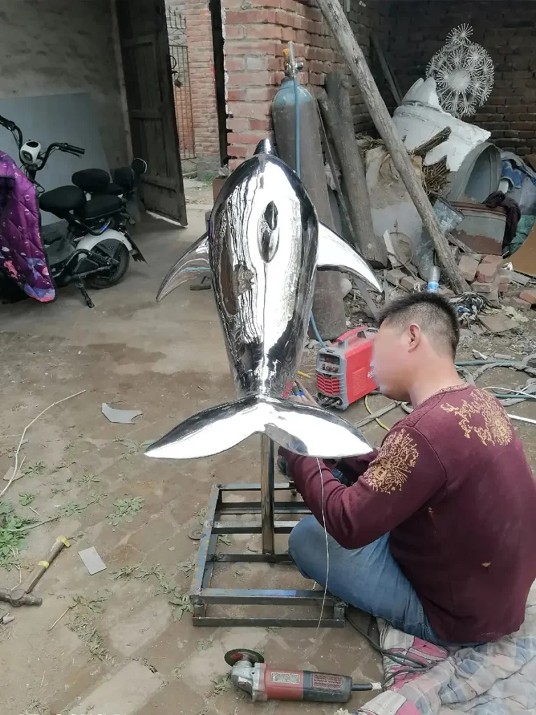 A skilled worker meticulously welding and grinding the base of a 316 marine-grade stainless steel dolphin sculpture. Proper fabrication of this specialized alloy ensures the contemporary pool statue remains structurally stable and totally chlorine-resistant for long-term outdoor exposure.