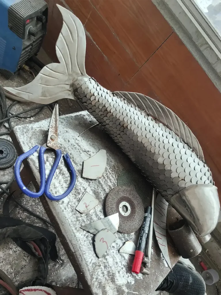 A metal fish sculpture mid-fabrication on a workshop table, surrounded by tools. The intricate detailing of the individual fish scales highlights the artisan craftsmanship required before the final polishing stage brings these dynamic aquatic animals to life.