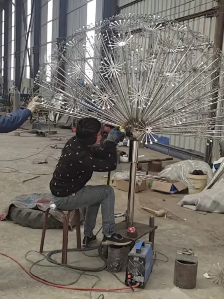An artisan meticulously welding the intricate, fine filaments of a giant stainless steel dandelion sculpture in a busy workshop. This highly detailed metal flower sculpture introduces a delicate softness into rigid environments, proving how expert craftsmanship transforms cold metal into a quiet, meditative presence.